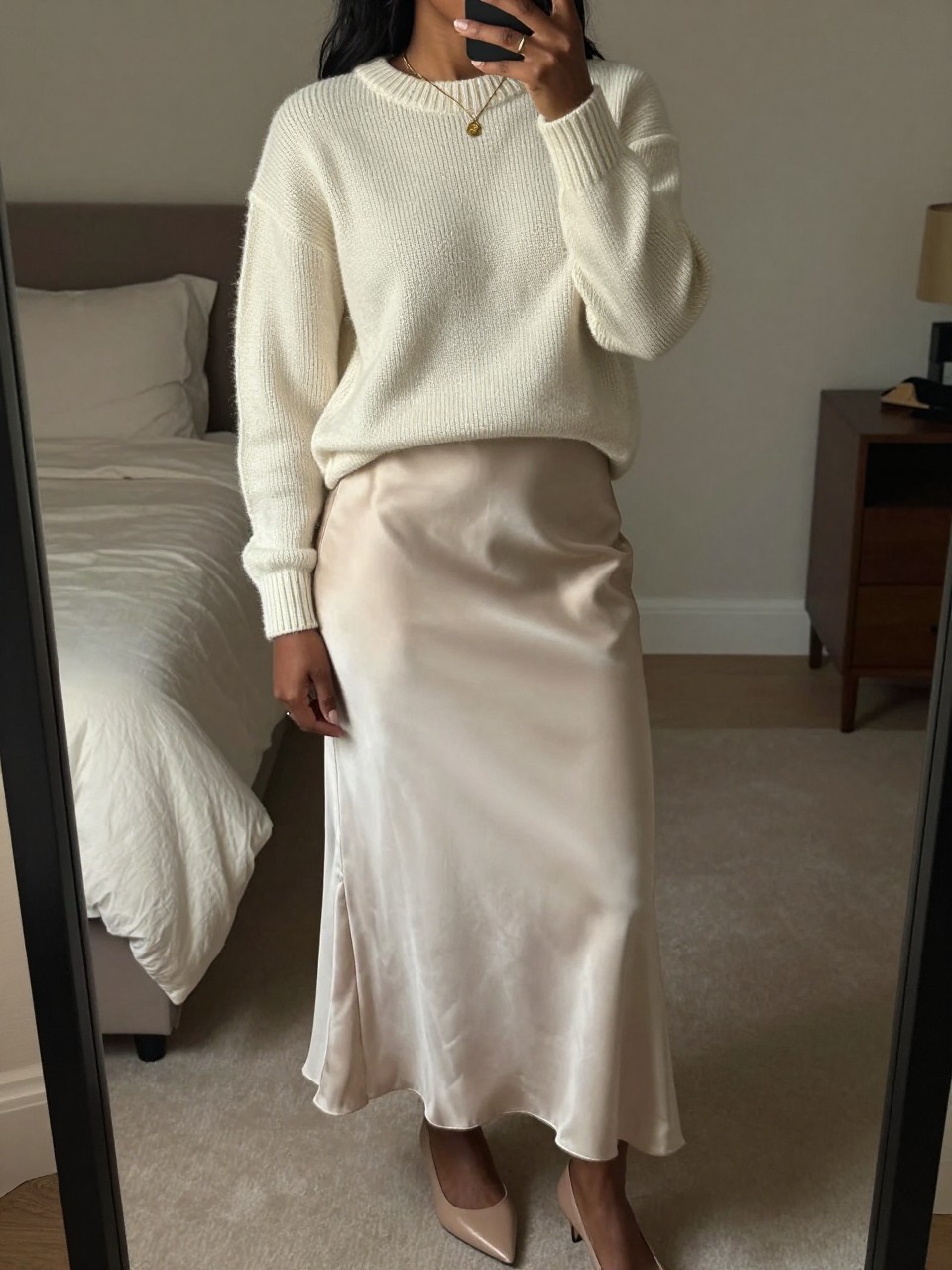 mirror selfie of a black woman wearing a cream cashmere crewneck sweater tucked into a cream satin maxi skirt creating a monochrome look, with nude pointed flats and minimal gold jewelry, standing in a sophisticated pose, cropped at NECK so face doesn't show, in a feminine-themed bedroom, warm moody lighting, iPhone photo quality.