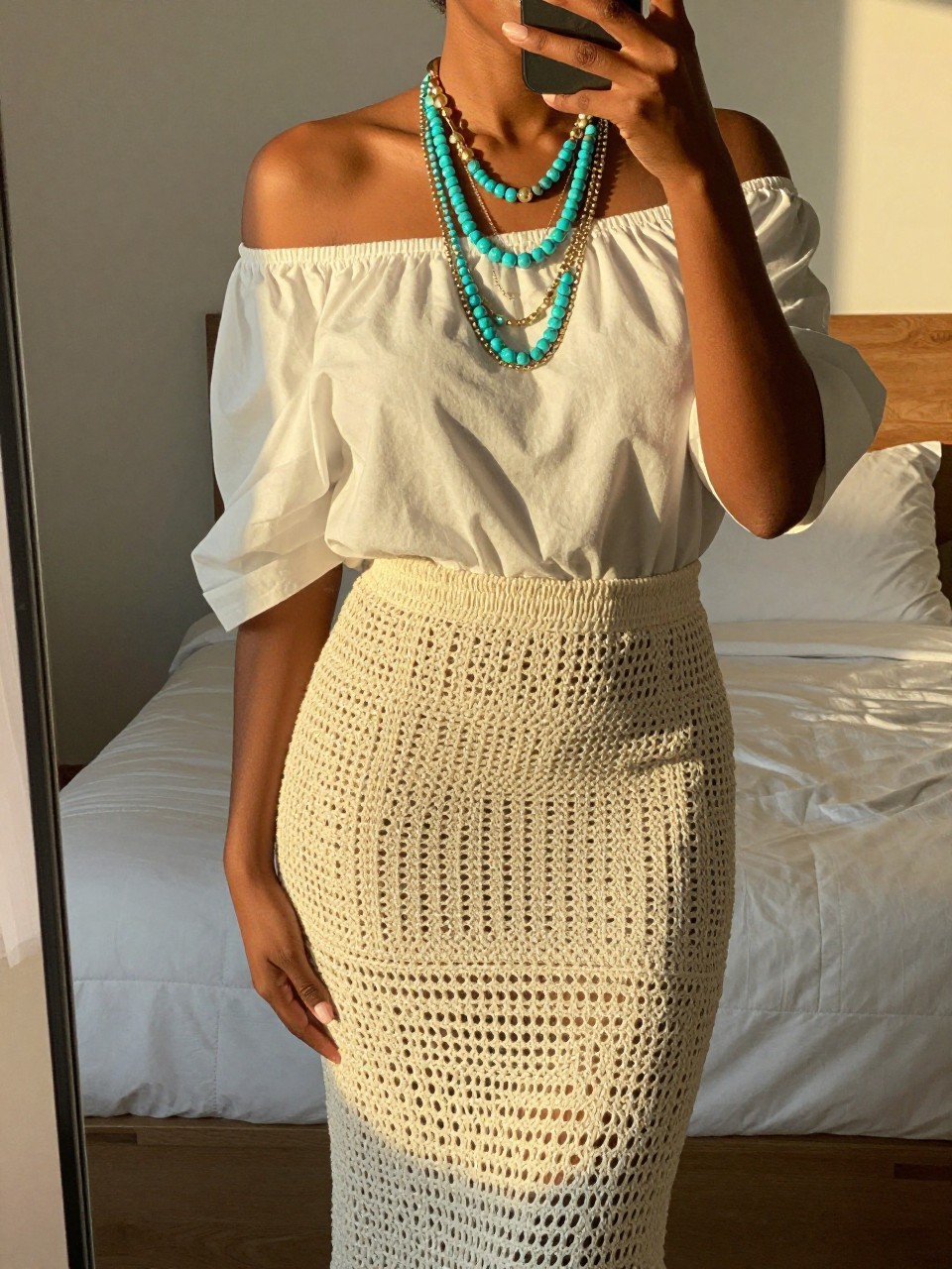 iPhone mirror selfie of a black woman wearing a white off-the-shoulder blouse with a natural crochet maxi skirt and layered turquoise and gold necklaces, one hand touching her necklaces, cropped at NECK so face doesn't show, in a feminine-themed bedroom, evening golden hour, iPhone photo quality.