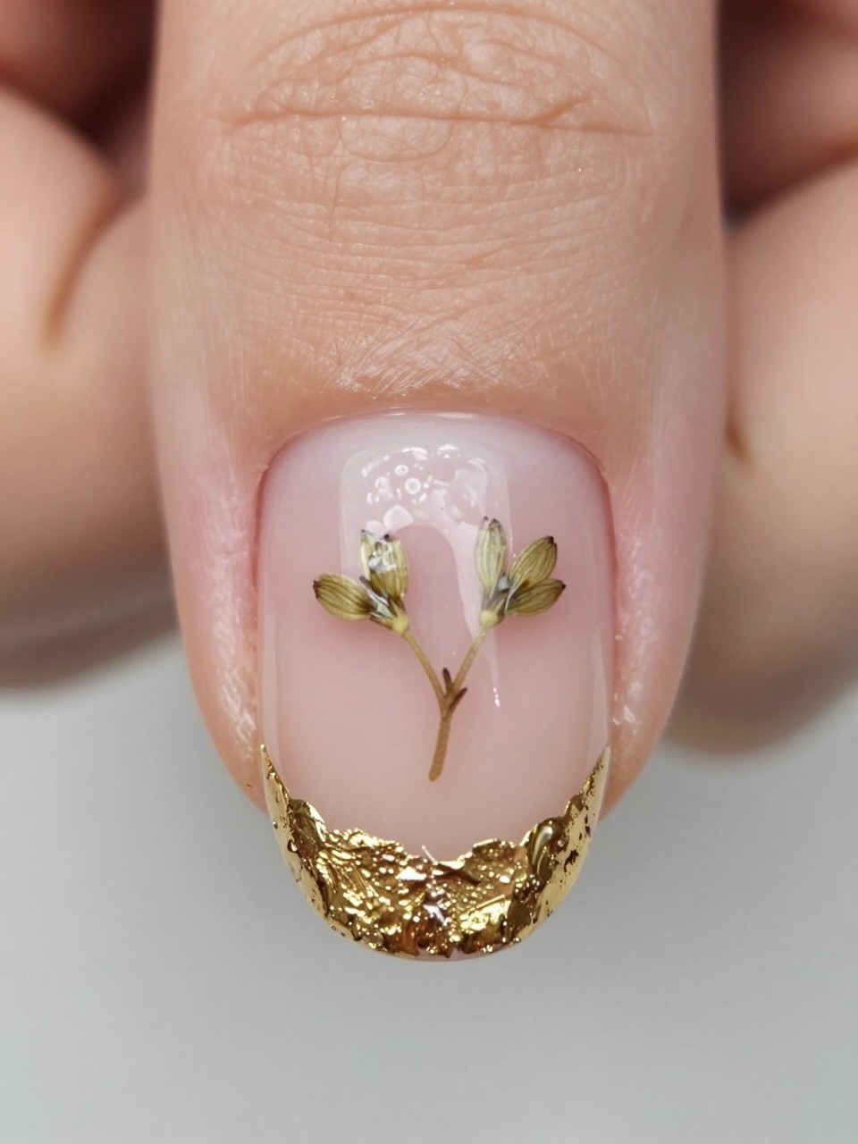 Macro, top-down view of a nail. The tip is made of crumbled gold leaf over a clear base. On the nude nail bed, a real pressed baby's breath sprig is encapsulated under gloss. The contrast is rich and natural.