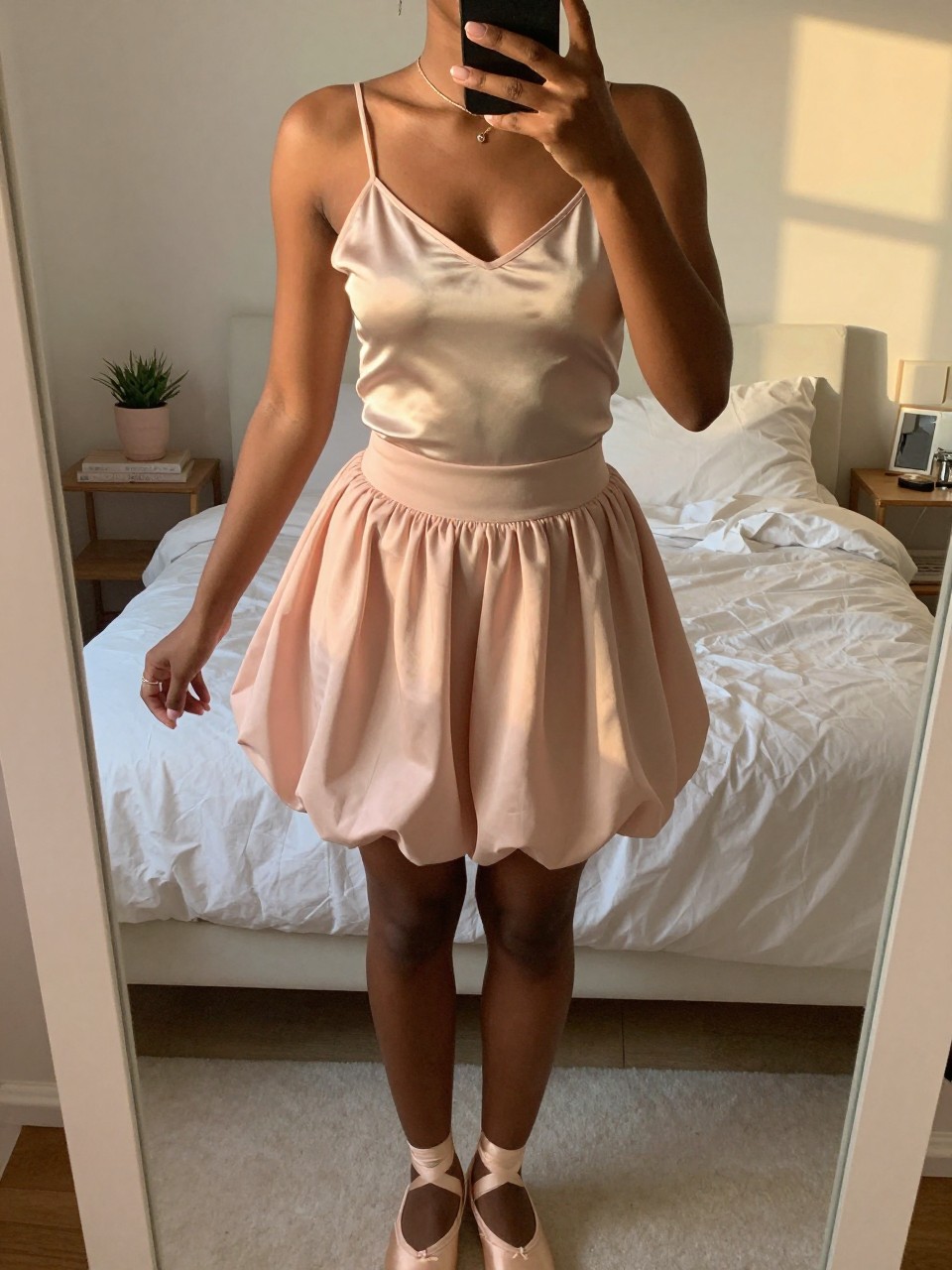 iPhone mirror selfie of a black woman in a champagne silk camisole and a blush pink bubble skirt with ballet flats, gently adjusting her necklace, cropped at NECK so face doesn't show, in a feminine-themed bedroom, evening golden hour, iPhone photo quality.