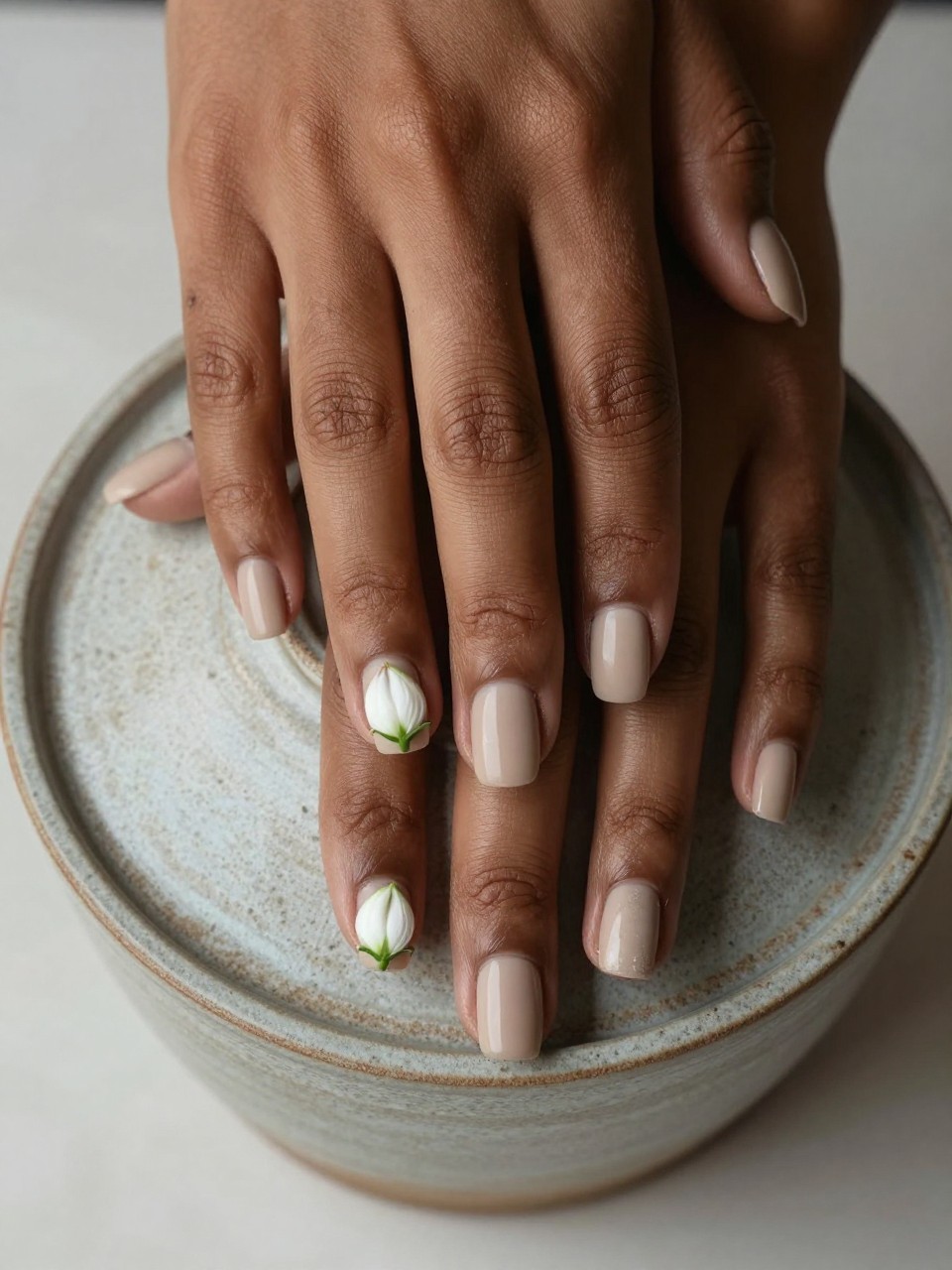 Overhead view of black feminine hands with short nails resting on a textured ceramic pot. The nails are a matte sandy beige. Two micro white buds with a hint of green are painted at the base of each nail. The look is organic and calm.