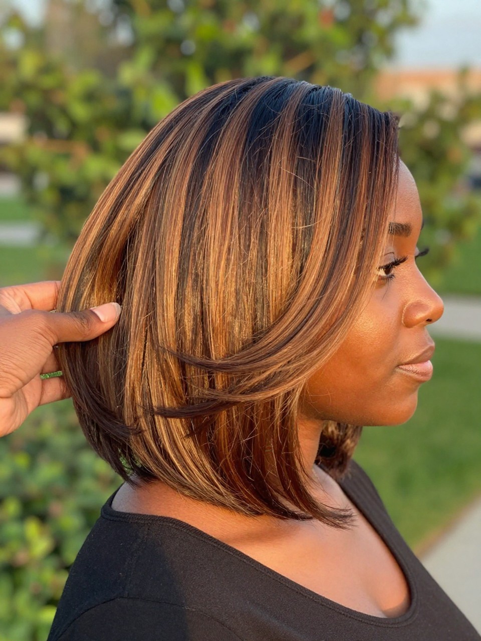 Photo of a black woman wearing a layered bob with caramel highlights, side profile view showing the dimension in the layers, setting is golden hour light outdoors, blurred greenery background, containing her hand gently touching a highlighted piece, iPhone photo quality.