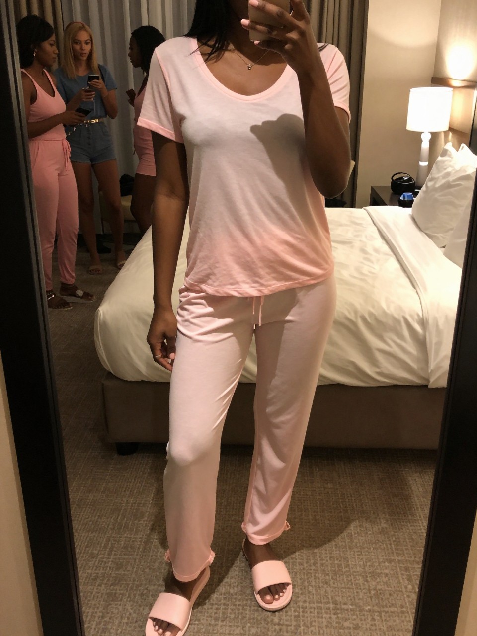 mirror selfie of a black woman in a pink lounge set and slides, standing in a hotel room with friends visible in the background, cropped at NECK so face doesn't show, in a feminine-themed bedroom, warm moody lighting, iPhone photo quality.