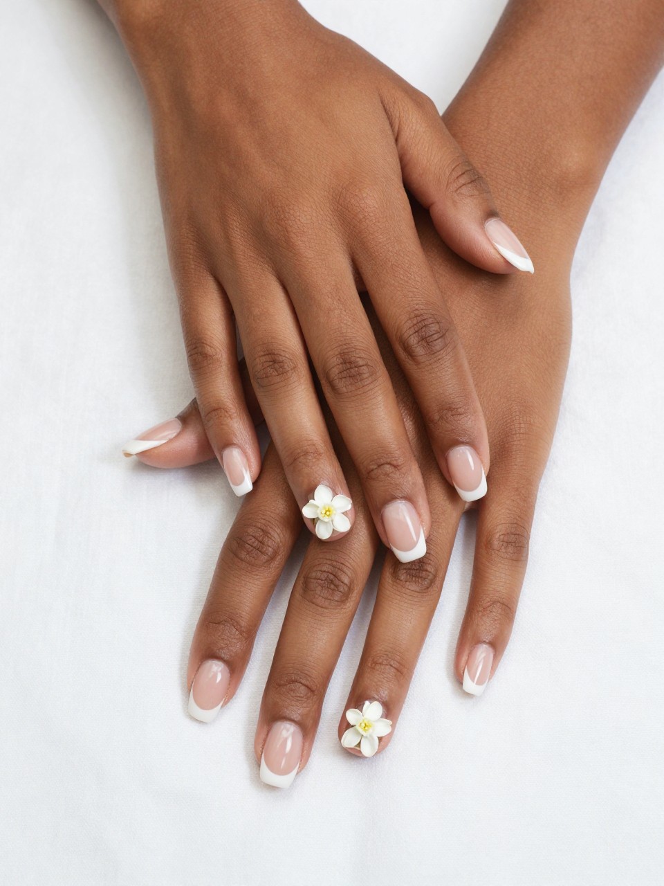 Overhead shot of black feminine hands with a classic French manicure, resting on a white cotton sheet. On the natural nail bed of each finger, a single micro white jasmine flower is painted near the tip. The composition is clean, balanced, and subtly detailed.