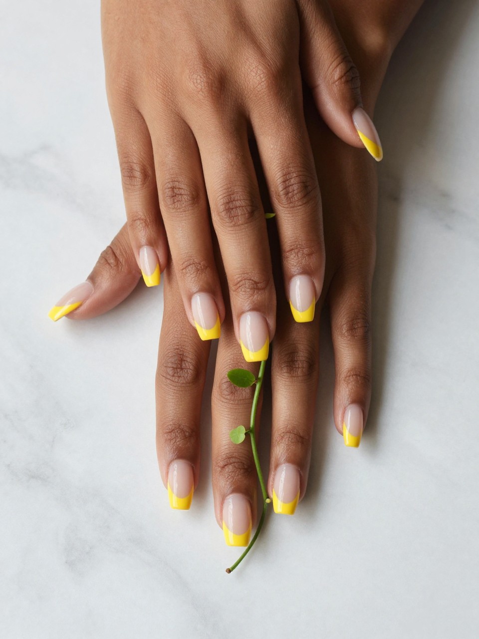 Overhead shot of black feminine hands with natural, long nails on a marble countertop. The nails have a daffodil yellow French tip. A single green vine with tiny leaves grows from the tip down the nail bed. The look is elegant and fresh.