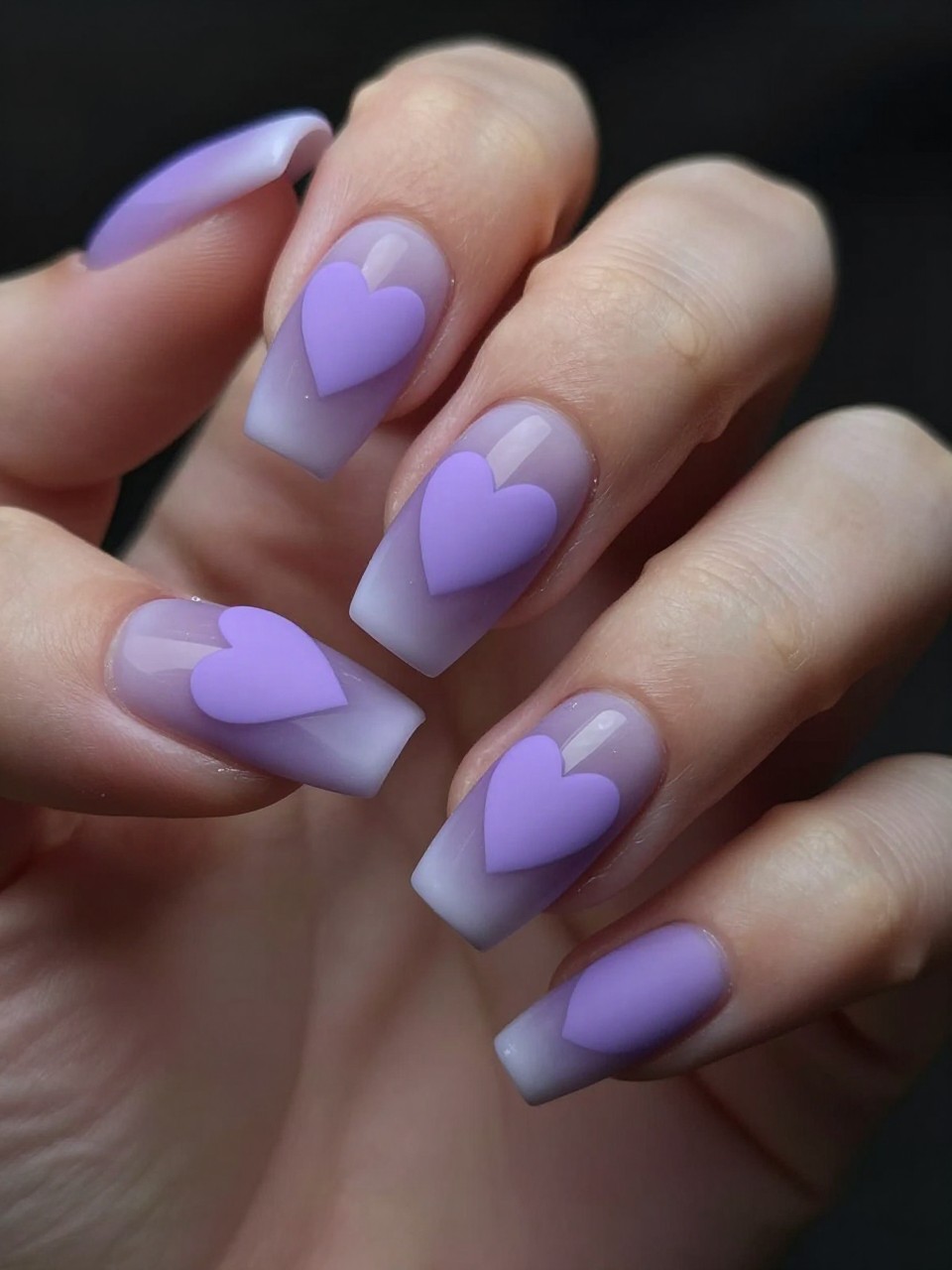Angled view of black feminine nails in moody, soft light. Each nail has a matte purple base with a soft white smoky halo. A solid lavender heart sits in the clear center. The effect is dreamy and focused.