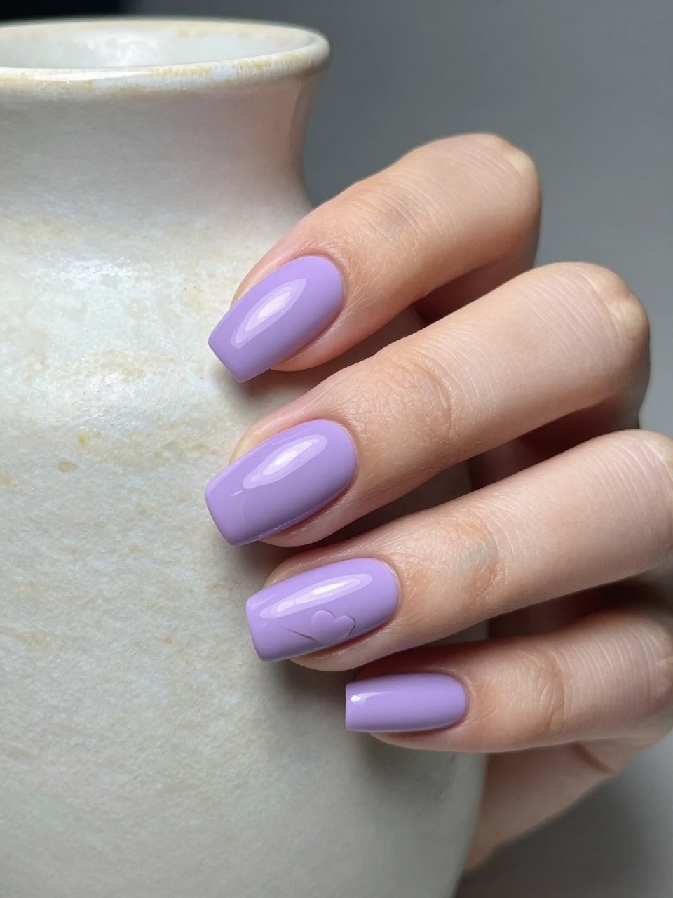 Side-angle shot of nails under a soft light source. The base is a matte wisteria purple. A small, glossy heart of the same color catches the light in the center of each nail. The hand rests on a textured ceramic vase.