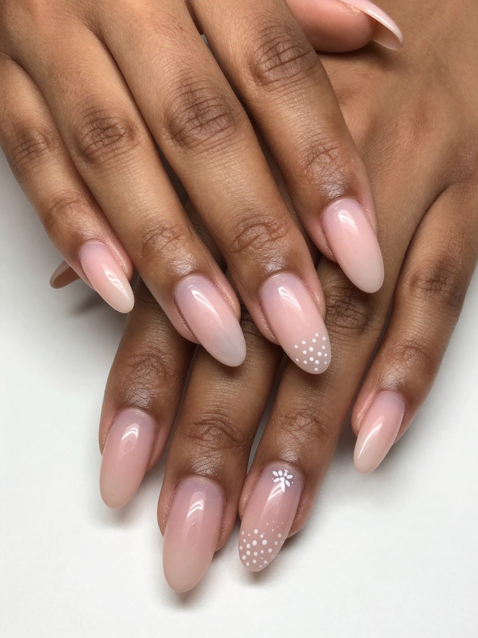 Angled view of black feminine hands with long nails. The nails show a soft blush ombré, deepest at the cuticles. A faint line of tiny white dot flowers sits right at the base where the color is strongest. The effect is ethereal and romantic.
