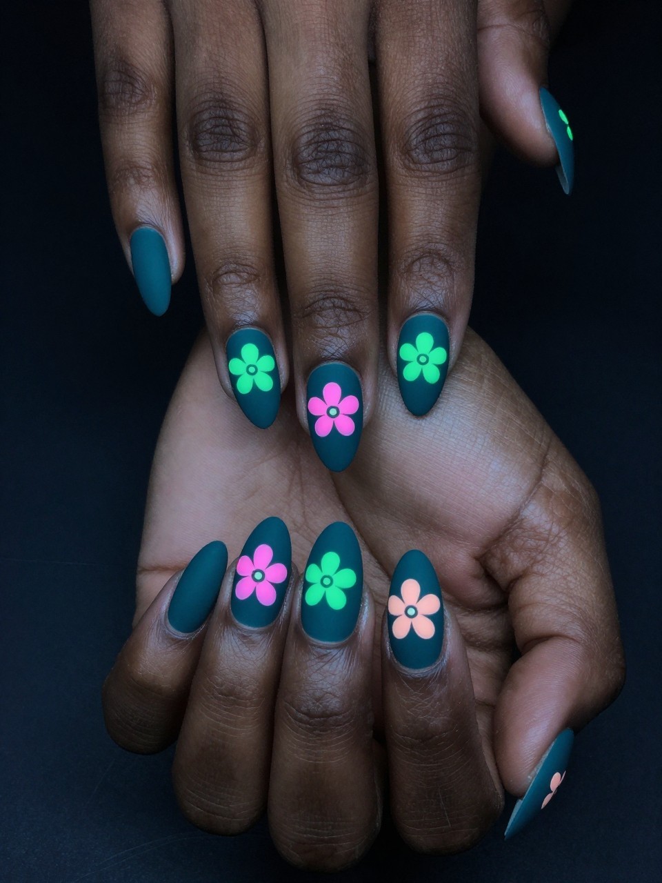 Top-down view of black feminine hands with almond nails against a dark, moody background. The nails are a matte deep teal. Graphic, simple floral shapes in neon pink and green are painted on each nail, appearing to glow under UV or black light. The look is modern and edgy.