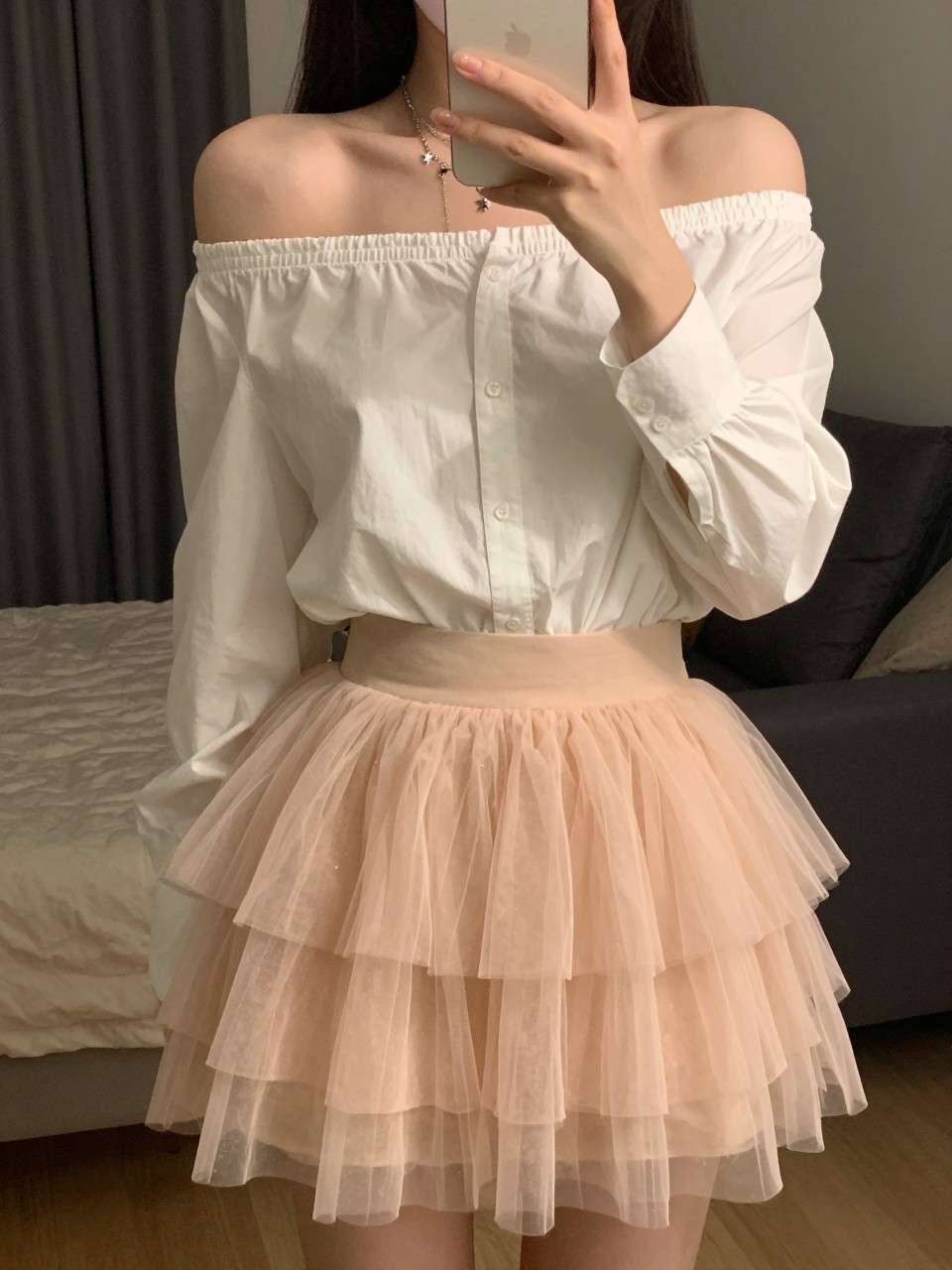 mirror selfie of a woman wearing a white off-the-shoulder blouse with a layered blush tulle fairy skirt and layered delicate star necklaces, one hand holding her phone at chest level, cropped at NECK so face doesn't show, in a city apartment, warm moody lighting, iPhone photo quality.