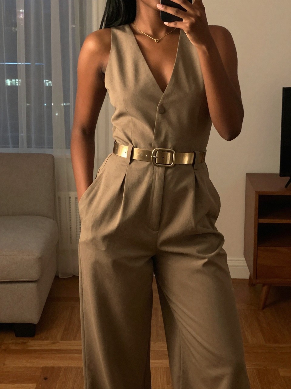 selfie of a black woman in a wide-leg jumpsuit with a metallic belt, posing with hands in pockets, cropped at NECK so face doesn't show, in a city apartment, warm moody lighting, iPhone photo quality.