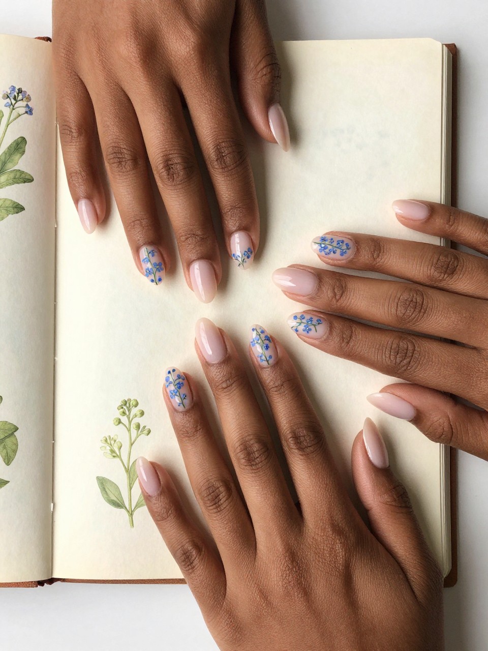 Bird's-eye view of black feminine hands with oval nails resting on a vintage botanical journal. The nails have a sheer blush base. On each nail design is a single, vertical sprig of tiny blue forget-me-nots is painted on the side of each nail. The mood is serene, orderly, and scholarly.