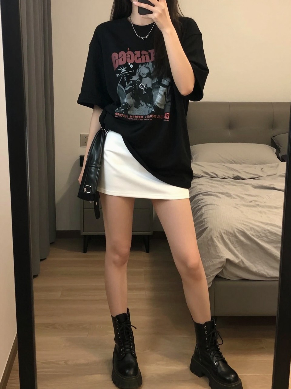 mirror selfie of a woman wearing an oversized black graphic tee loosely tucked into a white mini skirt with chunky combat boots, a leather jacket tied at her waist, standing in a confident power stance, cropped at NECK so face doesn't show, in a feminine-themed bedroom, warm moody lighting, iPhone photo quality.