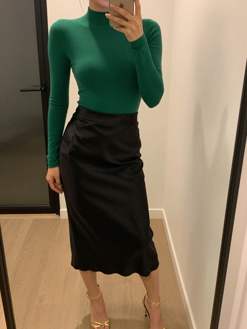 iPhone mirror selfie of a woman wearing an emerald green long-sleeve bodysuit tucked into a black satin midi skirt with gold strappy stiletto heels, one hand on her hip holding her phone, cropped at NECK so face doesn't show, in a city apartment, warm moody lighting, iPhone photo quality.