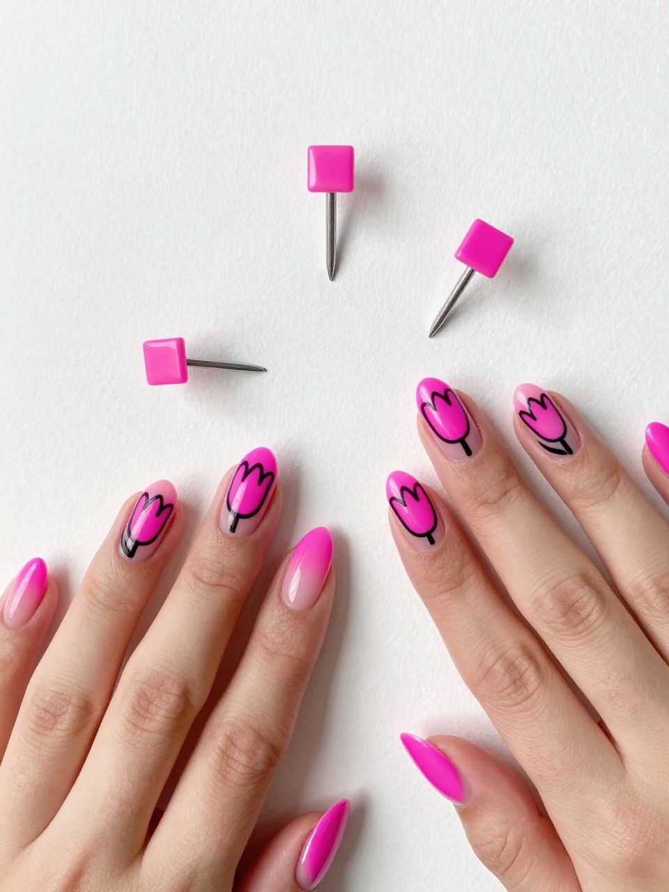 Overhead shot of hands with square nails against a white wall. The nails have neon pink French tips. A bold, black outline of a tulip is painted on each natural nail bed. The composition is graphic and eye-catching.
