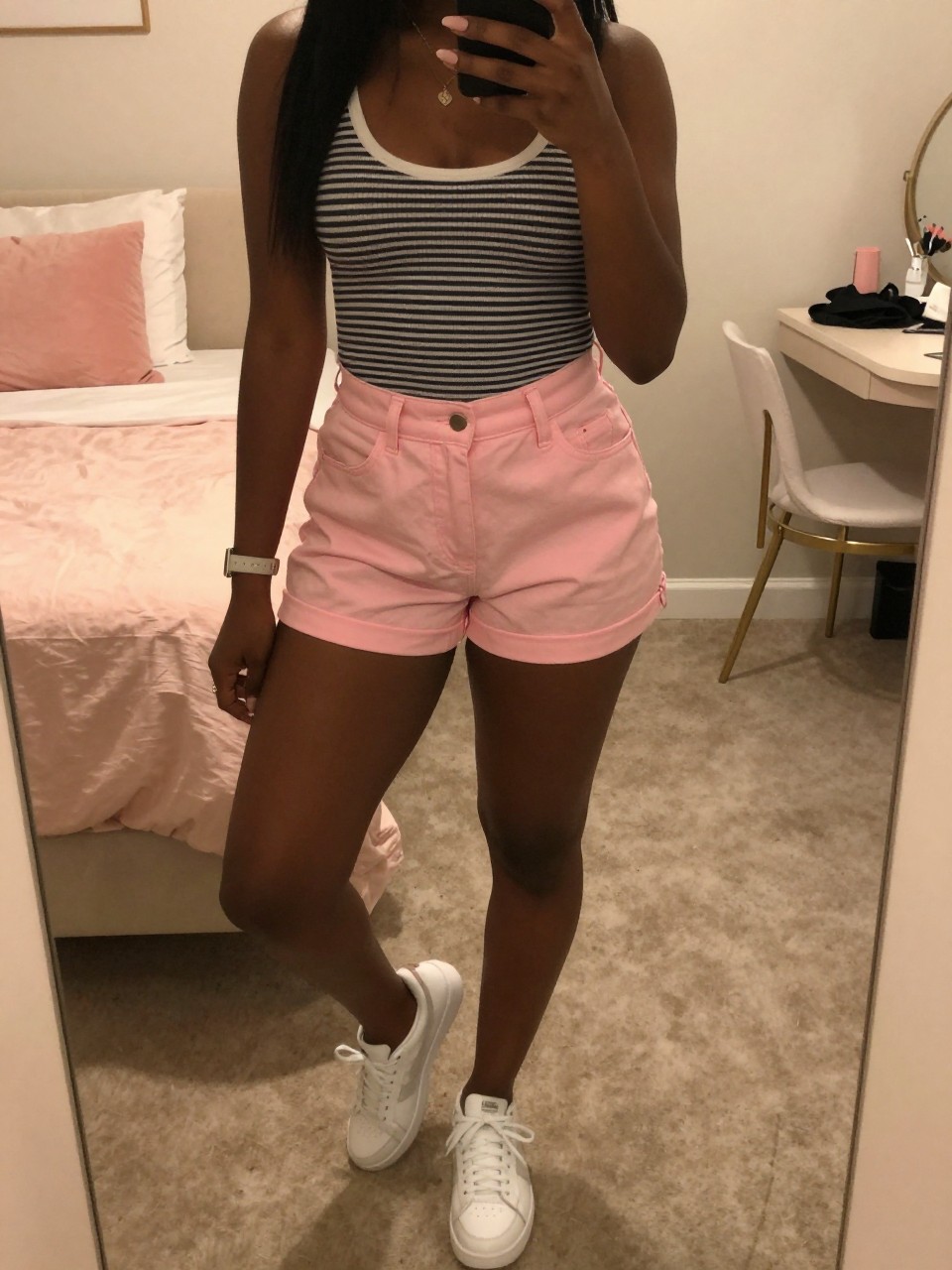 mirror selfie of a black woman in pink shorts and a striped top with white sneakers, standing with one foot forward, cropped at NECK so face doesn't show, in a feminine-themed bedroom, warm moody lighting, iPhone photo quality.