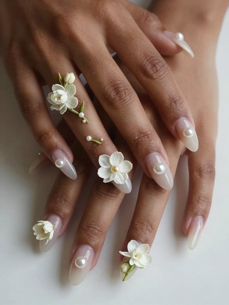 Close-up, bird's-eye view of black feminine hands with long nails. A vine of tiny white jasmine flowers trails across each sheer nail, each bloom centered with a micro pearl. The lighting is soft, making the pearls glint gently.