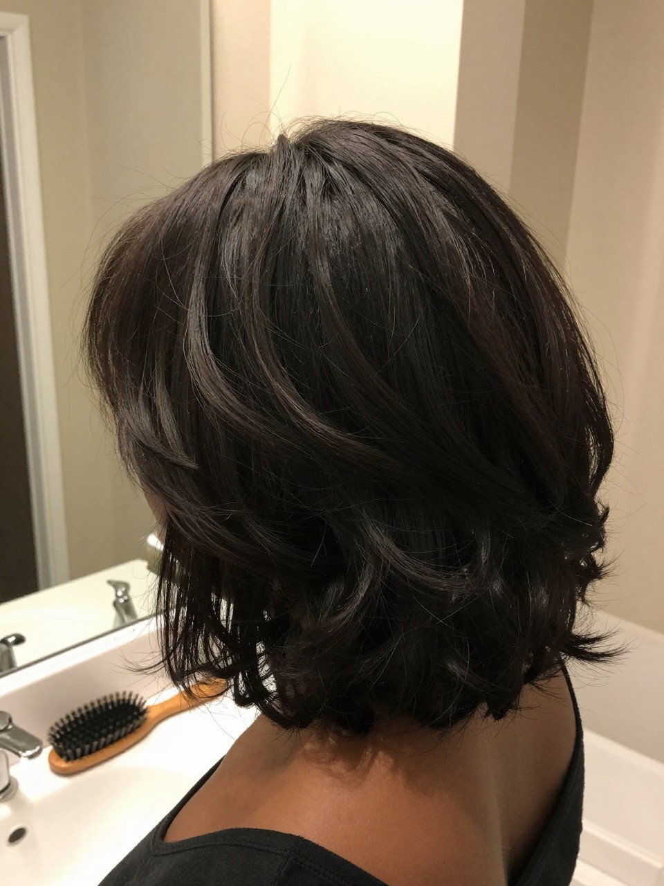 Photo of a black woman wearing a bouncy blowout bob, back-of-head view showing the layers and movement, setting is a bathroom vanity mirror with soft indoor lighting, containing a round brush on the counter, iPhone photo quality.