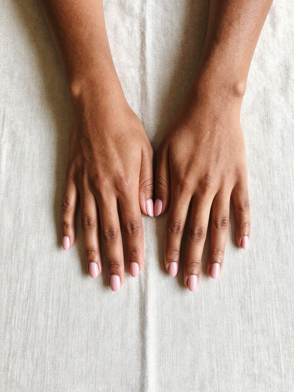 Overhead, top-down view of black feminine hands with short, neatly rounded nails resting palms down on a light beige linen tablecloth. The nails are painted a soft, dusty blush pink with a flawless matte finish. The lighting is bright and natural, showing off the clean lines and velvety texture.