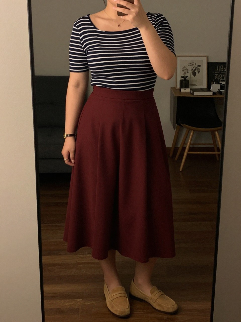 mirror selfie of a curvy woman wearing a navy striped boatneck top tucked into a maroon pleated midi skirt with tan loafers, one hand adjusting her collar, cropped at NECK so face doesn't show, in a city apartment, warm moody lighting, iPhone photo quality.