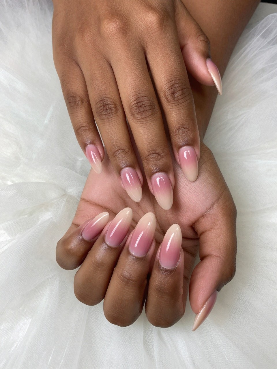Overhead, top-down view of black feminine hands with almond-shaped nails resting on a cloud of tulle fabric. The manicure is a seamless vertical ombré, deepest in a muted rose at the base and fading to a translucent beige at the free edge. The lighting is soft and ethereal, showing the perfect gradient.