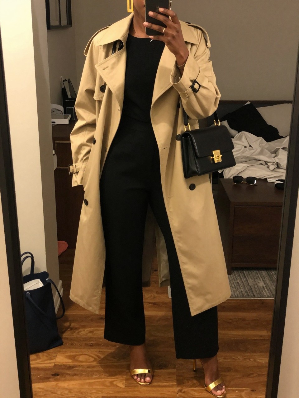 mirror selfie of a black woman wearing a black crepe wide-leg tailored jumpsuit with a warm beige light trench coat, metallic gold heeled sandals, bold clean gold statement earrings, and a black mini shoulder bag with gold hardware, holding a phone in front of a mirror, cropped at NECK so face doesn't show, with a natural casual selfie pose, in a city apartment, warm moody lighting, including small lived-in details like a tote bag on the floor, sunglasses on a dresser, or soft bedding in the background, latest iPhone photo quality.