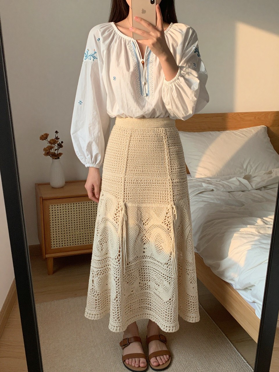 iPhone mirror selfie of a woman wearing a white embroidered peasant blouse with a natural crochet maxi skirt and flat leather sandals, one hand holding her phone at chest level, cropped at NECK so face doesn't show, in a feminine-themed bedroom, evening golden hour, iPhone photo quality.