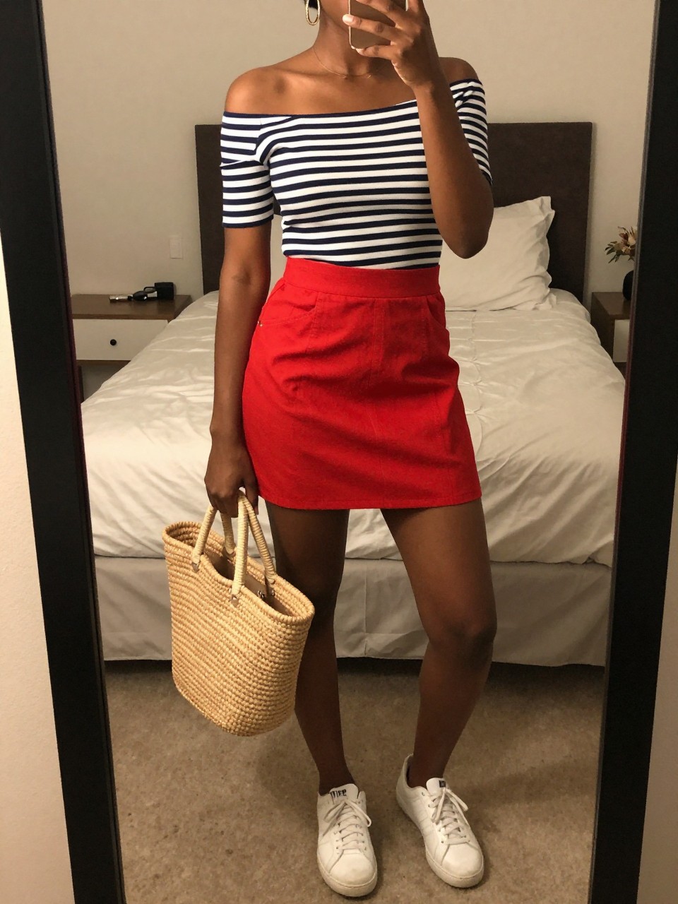 mirror selfie of a black woman wearing a navy and white striped boatneck top tucked into a bright red A-line mini skirt with white sneakers and medium gold hoop earrings, holding a straw tote bag, standing in a relaxed pose, cropped at NECK so face doesn't show, in a feminine-themed bedroom, warm moody lighting, iPhone photo quality.