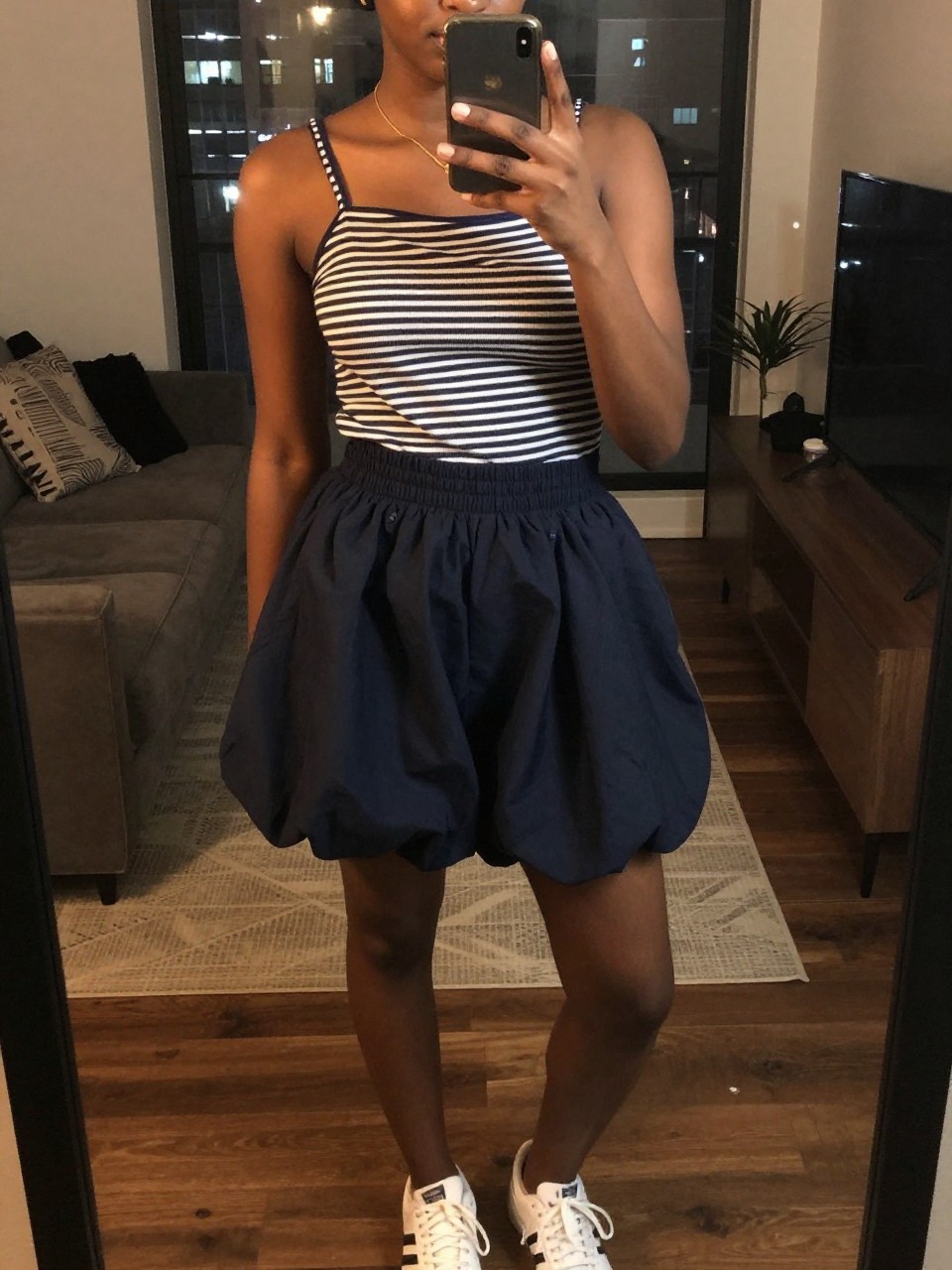 iPhone mirror selfie of a black woman in a striped boatneck top and a navy bubble skirt with white sneakers, holding her phone up to take a mirror pic, cropped at NECK so face doesn't show, in a city apartment, warm moody lighting, iPhone photo quality.