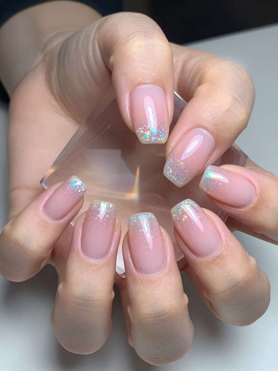 Close-up, bird's-eye view of hands under a prism catching light. The oval nails have a gradient from a solid ballet pink base to tips infused with fine, scattered holographic glitter. The light breaks into tiny rainbows on the holographic tips.