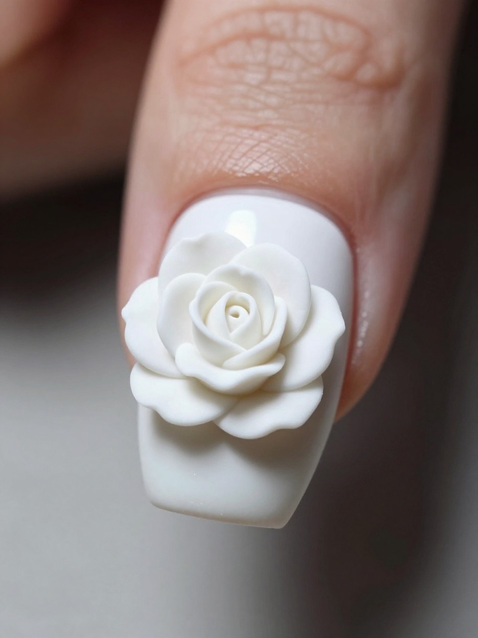 Side-angle, macro shot of a nail under soft light. The nail is glossy white. A single, intricately sculpted white rose with raised petals sits in the center, casting a soft shadow. The look is minimalist yet deeply luxurious.