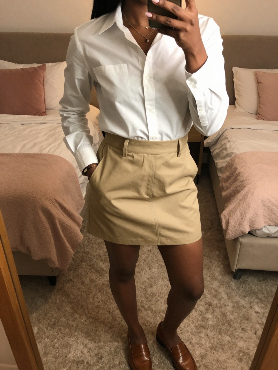 mirror selfie of a black woman wearing a white button-down shirt tucked into a khaki tennis skirt with brown leather loafers, one hand in her pocket, cropped at NECK so face doesn't show, in a feminine-themed bedroom, warm moody lighting, iPhone photo quality.