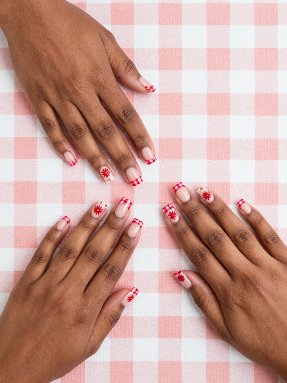 Overhead view of black feminine hands with short, square nails on a checkered tablecloth. The French tips are red and white checkered. On the nude base of each nail, a single red daisy is painted. The look is coordinated and fun.