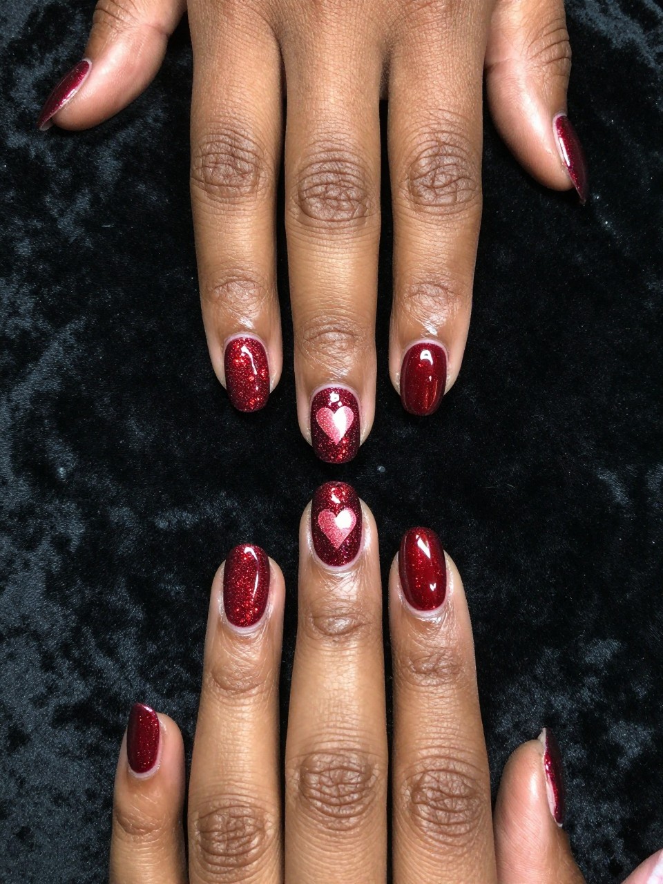 Overhead, top-down view of black feminine hands with medium coffin nails resting on a black velvet cloth. The nails feature a deep red cat eye polish. The magnetic glitter is intensely pulled into a bright, defined heart shape in the center of each nail. Dramatic lighting catches the shimmering heart against the darker base.