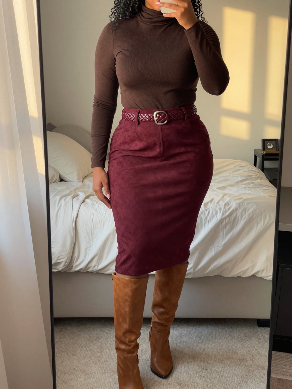 mirror selfie of a curvy black woman wearing a chocolate brown turtleneck tucked into a maroon suede skirt with a braided belt and cognac knee-high boots, one hand resting on her belt, cropped at NECK so face doesn't show, in a feminine-themed bedroom, evening golden hour, iPhone photo quality.