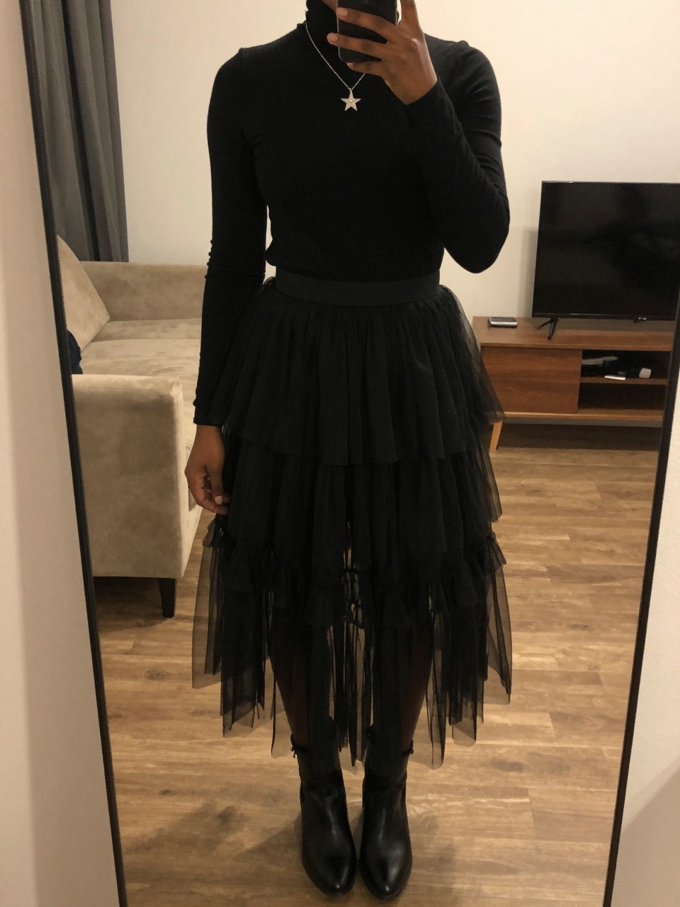 mirror selfie of a black woman wearing a black turtleneck tucked into a black layered tulle fairy skirt with a silver star necklace and black ankle boots, one hand touching her necklace, cropped at NECK so face doesn't show, in a city apartment, warm moody lighting, iPhone photo quality.