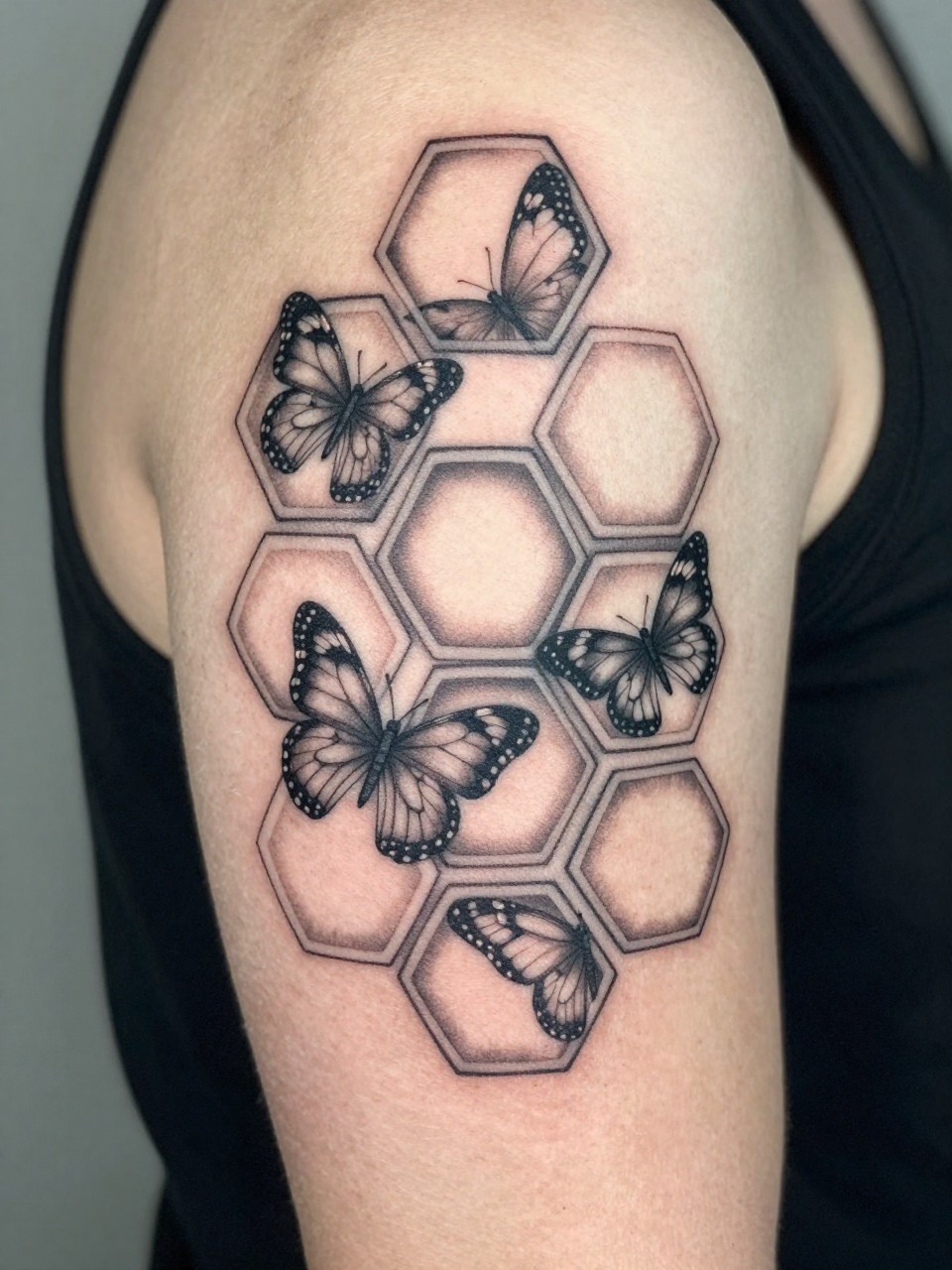 Detailed view of an upper arm tattoo: a honeycomb geometric pattern where select hexagons contain fragments of butterfly wings. Clean, sharp lines, black and grey ink."