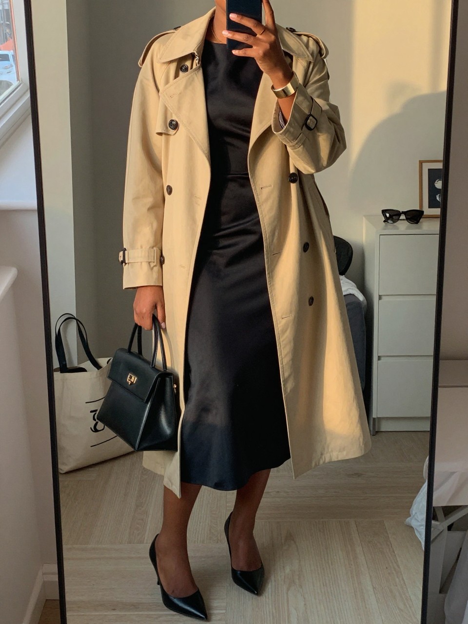 mirror selfie of a black woman wearing a smooth black crepe midi dress with a warm beige light trench coat, black pointed-toe heels, a structured black handbag, and a clean gold cuff bracelet, holding a phone in front of a mirror, cropped at NECK so face doesn't show, with a natural casual selfie pose, in a city apartment, evening golden hour, including small lived-in details like a tote bag on the floor, sunglasses on a dresser, or soft bedding in the background, latest iPhone photo quality.