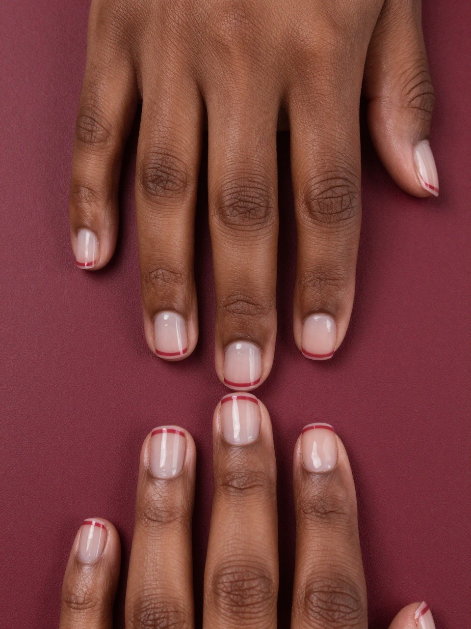 Close-up, top-down view of black feminine hands with short, square nails on a deep red journal. The base is a matte burgundy. A thin, glossy stripe runs straight down the middle of each nail, reflecting the light. The look is precise and rich.