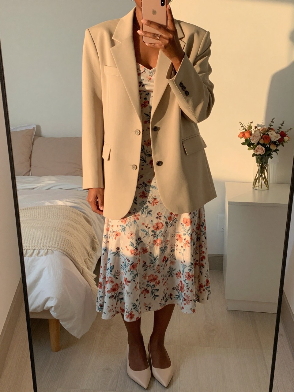 iPhone mirror selfie of a black woman in an oversized beige blazer and a soft floral midi skirt with pointed flats, standing with her phone in hand, cropped at NECK so face doesn't show, in a feminine-themed bedroom, evening golden hour, iPhone photo quality.