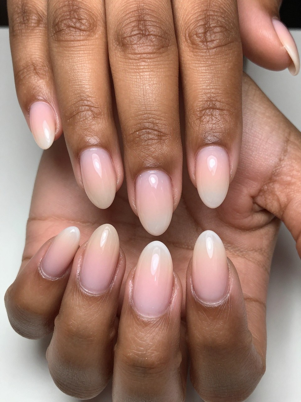 Extreme close-up, top-down view of black feminine hands with natural, medium almond nails. The ombré is incredibly subtle, fading from a tan nude at the cuticles to a translucent pink at the very tips. Clean, bright studio lighting reveals the masterful, subtle blend.