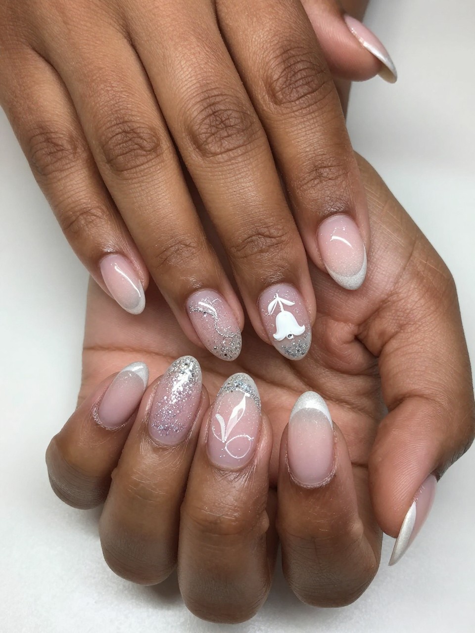 Close-up, top-down view of black feminine hands with oval nails. The French tips are a sparkling glitter fade and on the nail design, there is a single, small white bell flower designed on the nails glitter line. The mood is ethereal and festive.