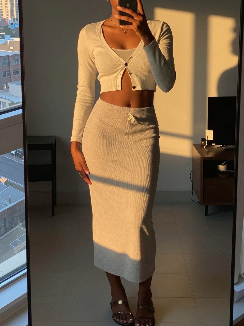 mirror selfie of a black woman in a long skirt and cropped cardigan over a tank with sandals, holding phone at waist, cropped at NECK so face doesn't show, in a city apartment, evening golden hour, iPhone photo quality.