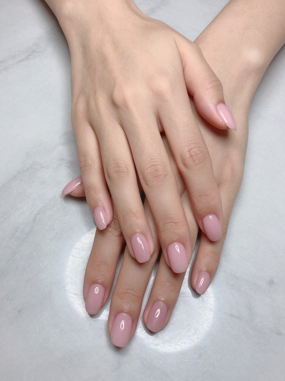Bird's-eye view of hands posed elegantly on a sleek, pale grey marble surface. The nails are a medium coffin shape, painted a soft pink and coated with a glossy, pearlescent top coat that gives a sugary glaze effect. The lighting reflects perfectly off the shiny surface.