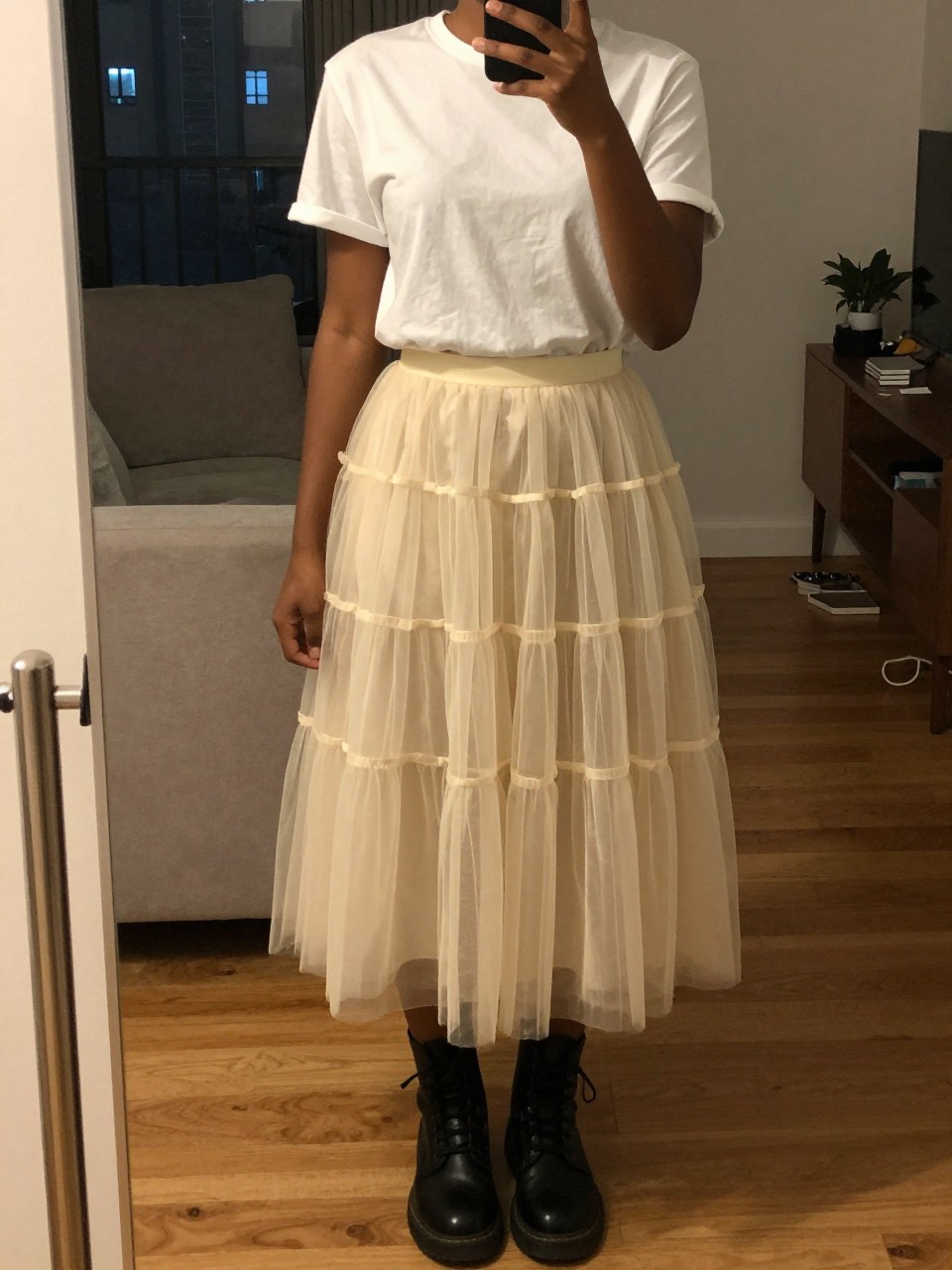 mirror selfie of a black woman wearing a simple white t-shirt with a layered cream tulle fairy skirt and black combat boots, one hand holding her phone at her side, cropped at NECK so face doesn't show, in a city apartment, warm moody lighting, iPhone photo quality.