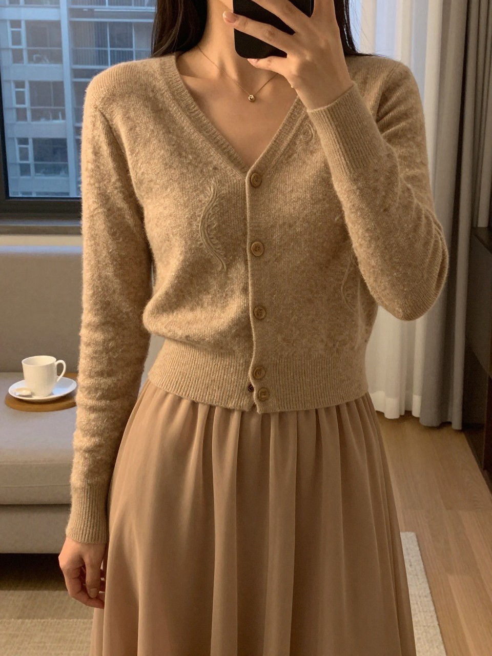 selfie of a woman in a cashmere set and chiffon skirt, cardigan buttoned, cropped at NECK so face doesn't show, in a city apartment, warm moody lighting, iPhone photo quality.
