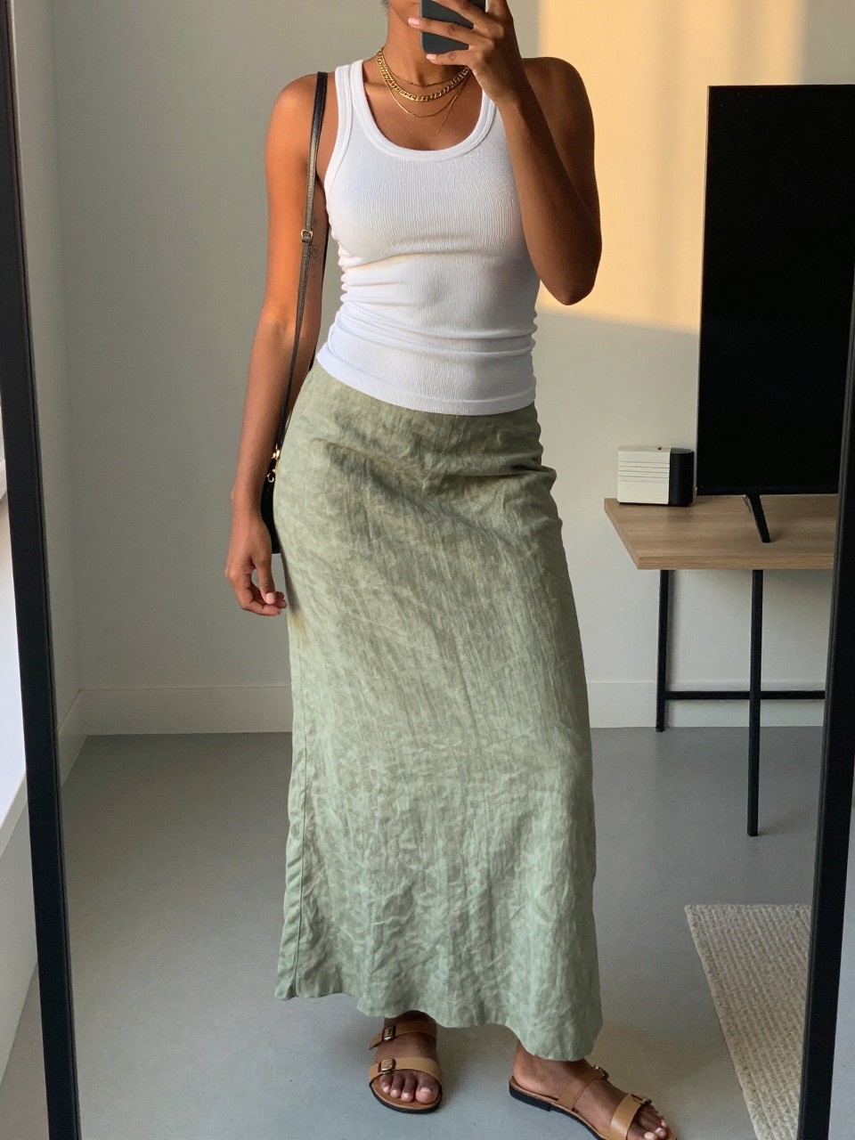 mirror selfie of a black woman wearing a white fitted ribbed tank top tucked into a sage green linen maxi skirt with layered gold necklaces and flat tan leather sandals, holding a small crossbody bag, standing in a relaxed minimalist pose, cropped at NECK so face doesn't show, in a city apartment, evening golden hour, iPhone photo quality.