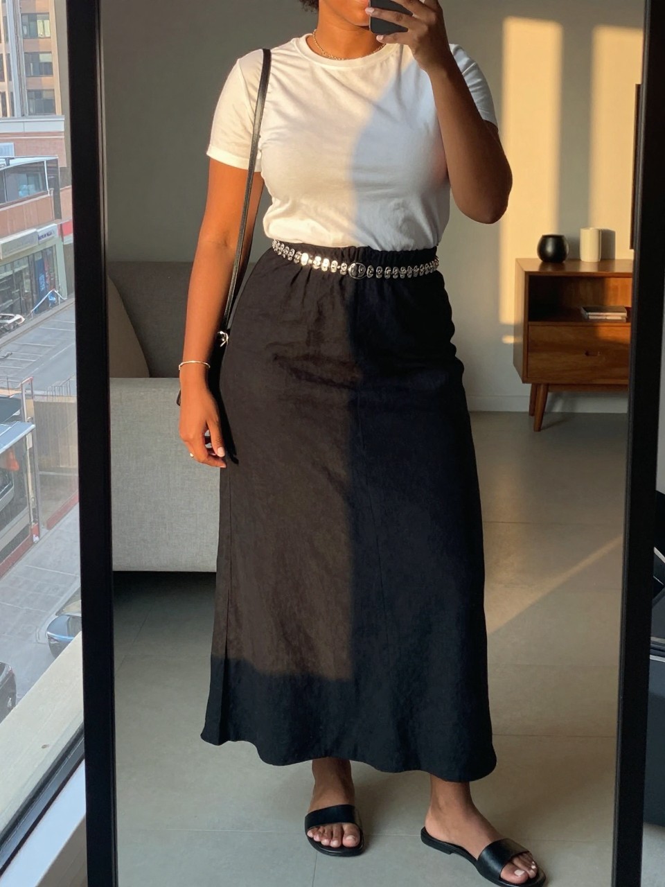 mirror selfie of a curvy black woman wearing a white crewneck t-shirt tucked into a black linen maxi skirt with a silver statement belt at her waist and black leather flat sandals, holding a black crossbody bag, standing in a casual elegant pose, cropped at NECK so face doesn't show, in a city apartment, evening golden hour, iPhone photo quality.