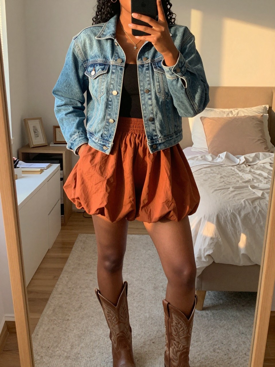 iPhone mirror selfie of a black woman in a denim jacket and a rust bubble skirt with cowboy boots, one hand in her jacket pocket, cropped at NECK so face doesn't show, in a feminine-themed bedroom, evening golden hour, iPhone photo quality.