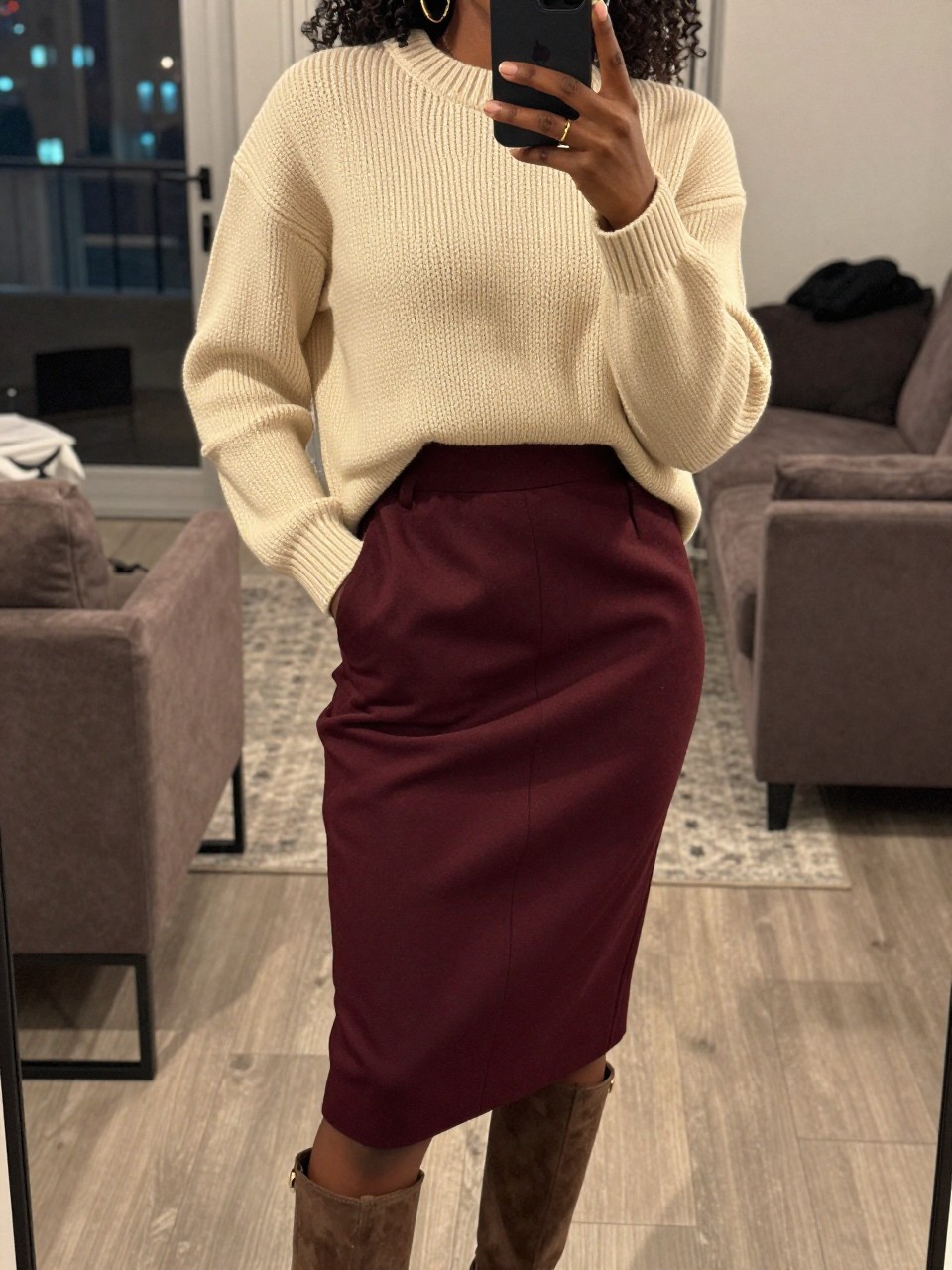mirror selfie of a black woman wearing a cream chunky sweater tucked into a maroon wool pencil skirt with gold hoops and suede boots, one hand holding a phone the other tucked into her coat pocket, cropped at NECK so face doesn't show, in a city apartment, warm moody lighting, iPhone photo quality.